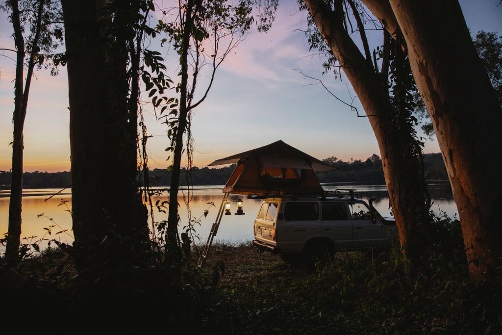 Picturesque scenery of camping tent placed on SUV car parked on coast of peaceful lake located in woods against amazing sunset sky
